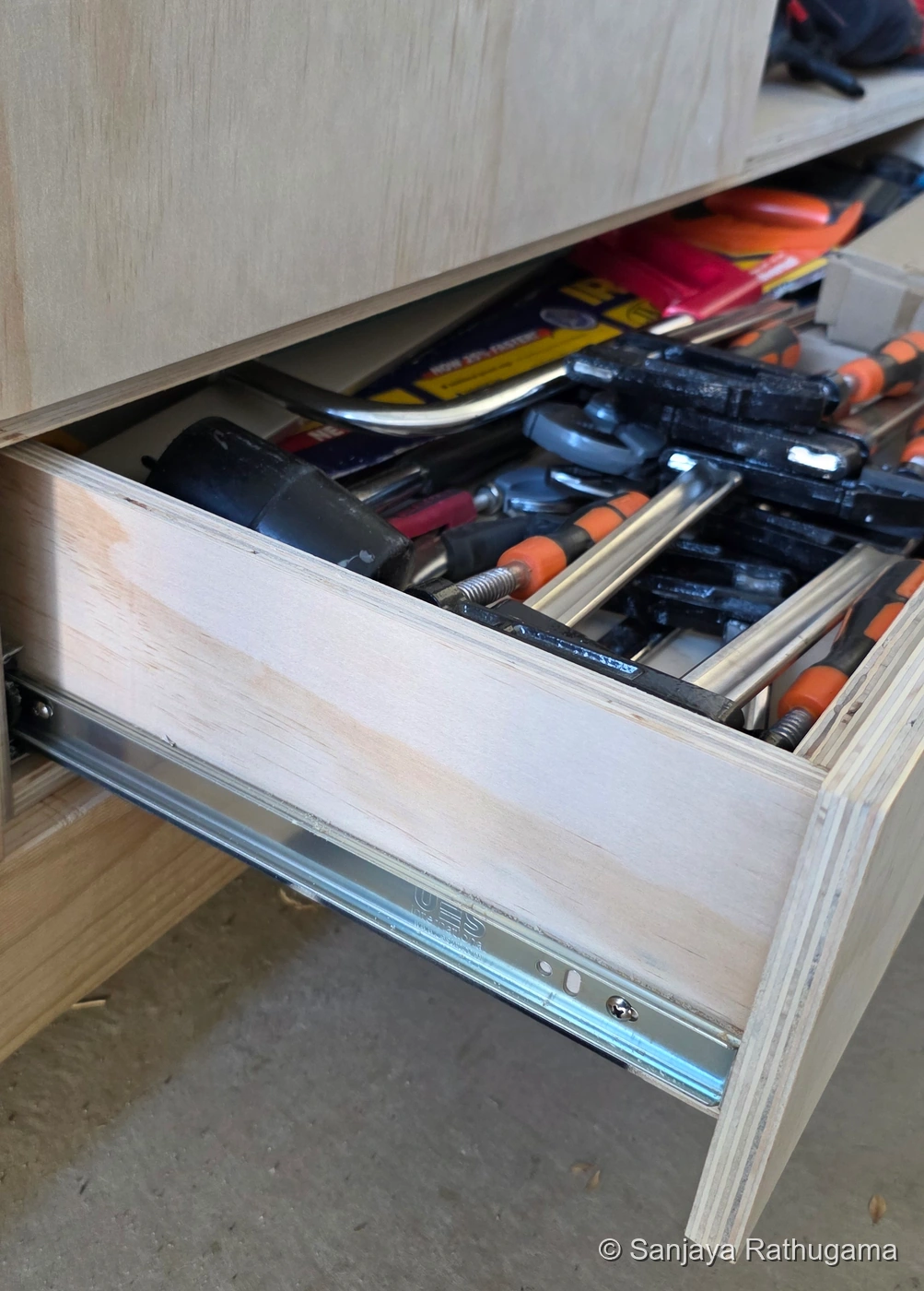 An open plywood drawer storing clamps and hand tools, with its telescopic runner and separate fascia visible